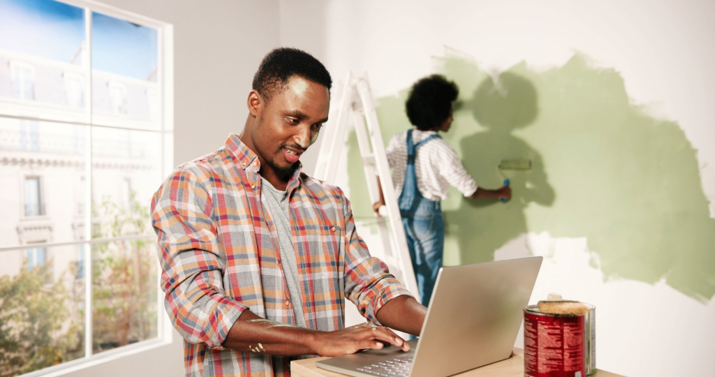 A man using a laptop while another man paints a wall